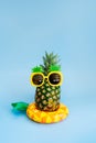 Funny pineapple in a sunglass of pineapple shape on the yellow inflatable ring on blue vertical background. Tropical style summer