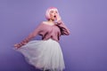 Funny pink-haired girl in periwig posing in studio. Adorable female model wears white skirt and bright peruke enjoying