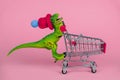 Plastic toy dinosaur wearing tiny knitted hat and driving empty shopping trolley on a pink background ,shopping concept