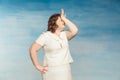 Funny plump woman in a white dress demonstratively suffers by hitting himself on the forehead on a blue background