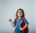 Funny portrait of happy little girl in glasses hugging a book. Child with backpack laughing and pointing with finger up Royalty Free Stock Photo