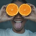 Funny portrait of young woman showing tongue out while covering her eyes with halves of orange