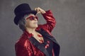 Happy youthful looking senior woman in top hat, cool glasses and sequin jacket posing in studio