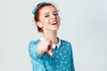 Funny redhead girl in light blue dress having, pointing finger at camera and toothy smile, focus on her face.