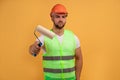 Funny repairman wears hardhat, apron, holds paint roller in hand for painting wall in yellow color. Craftsman uses special tool,