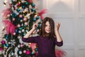 Funny Seven-year-old dark-haired girl in purple dress stands near the elegant Christmas tree indoors and looking to camera