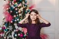 Funny Seven-year-old dark-haired girl in purple dress stands near the elegant Christmas tree indoors and looking to camera