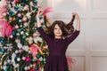 Seven-year-old dark-haired girl in purple dress stands near the elegant Christmas tree indoors and looking to camera