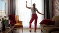 Funny shot of happy woman wearing pajamas and dancing against big window in living room. People relaxing at home, having fun,