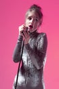 Funny Singing Caucasian Girl In Gray Reflective Dress Posing With Microphone Indoors On Pink Background