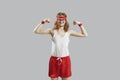 Funny skinny man in tank top and shorts showing weak muscles isolated on gray background