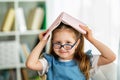 Funny smiling little girl with glasses and book on her head is sitting at table Royalty Free Stock Photo