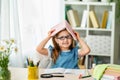 Funny smiling little girl with glasses and a book on her head sitting at table Royalty Free Stock Photo