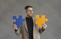 Funny smiling male office worker, makes up two colored puzzle pieces standing on a gray background.