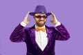 Funny smiling man in velvet jacket adjusts his hat while standing on purple studio background.