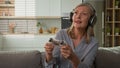 Funny smiling mature caucasian woman playing video game console in headphones using joystick controller at home on couch Royalty Free Stock Photo