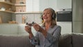 Funny smiling mature caucasian woman playing video game console in headphones using joystick controller at home on couch Royalty Free Stock Photo