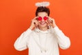 Funny smiling young man in casual hoodie with aureole overhead holding toy hearts near his eyes, pretending to be saint valentine