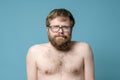 Funny man in glasses with a shaggy beard and an overgrown hairstyle, with a naked torso, stares into camera, squinting