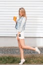 Funny stylish young woman in trendy sports dress in blue denim jacket in white sneakers with an orange cup with a cocktail