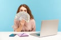 Funny surprised woman employee sitting at workplace with laptop and peeking out of dollar banknotes