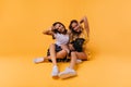 Funny tanned girls posing on the floor with dog. Studio shot of stunning white sisters isolated on yellow background Royalty Free Stock Photo