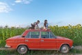 Funny teen children in casual clothes playing together on the red retro car among the sunflowers field, girl sitting on Royalty Free Stock Photo