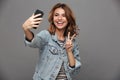 Funny teen girl in jeans jacket showing peace gesture while taking selfie on smartphone Royalty Free Stock Photo
