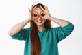 Funny teen girl with long ginger hair, look through finger binoculars, make glasses with hands and smiling happy Royalty Free Stock Photo