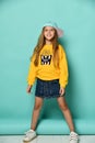 Funny teenage girl in a baseball cap and a yellow hoodie and a denim miniskirt posing in full growth on a blue background. Royalty Free Stock Photo