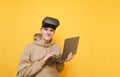 Funny teenager in casual clothes stands on a yellow background with a laptop in his hands with VR glasses on his head, looks into