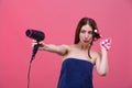 A funny girl is in a towel and with hair curlers, holds a hair dryer and directs it in front of her. Pink background.