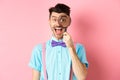 Funny young man in bow-tie look through magnifying glass and showing tongue, smiling happy, having fun on pink Royalty Free Stock Photo