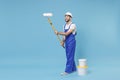 Funny young man in coveralls protective helmet hardhat hold paint roller, bucket isolated on pastel blue background Royalty Free Stock Photo