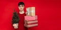 Funny young man with a frowning face holds a Christmas tree and gifts on a red background