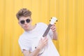 Funny young man in glasses and a white T-shirt plays ukulele on a yellow background