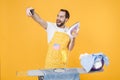 Funny young man househusband in apron ironing clean clothes on board doing housework isolated on yellow wall background