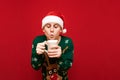 Funny young man in santa hat and green sweater stands with cup of hot drink in hands on red background. Serious guy in christmas Royalty Free Stock Photo