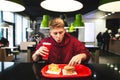 Funny young man sitting in a fast-food restaurant, holding a glass of drink and taking a burger from the tray Royalty Free Stock Photo