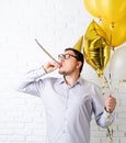 Funny young man wearing birthday hat and holding balloons blowing noisemaker celebrating birthday