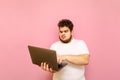 Funny young overweight man uses a laptop on a pink background and with a concentrated face looks at the computer screen. Fat man Royalty Free Stock Photo