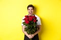 Funny young salesman in black apron holding bouquet of roses, florist laughing and standing over yellow background