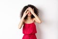 Funny young woman with curly hair, wearing red dress, covering eyes with hands but peeking through fingers and smiling Royalty Free Stock Photo