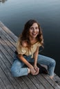 Funny young woman with a cute smile in blue jeans and yellow top t-shirts sits on the wooden pier by the lake. Summer weekends Royalty Free Stock Photo