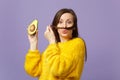 Funny young woman in fur sweater keeping hair like mustache, holding half of fresh ripe avocado isolated on violet