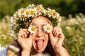 A funny young woman in a wreath of daisies is laughing and holding daisies in front of her eyes with her tongue hanging out. On a Royalty Free Stock Photo