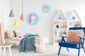 Furry pink pillow on a vibrant blue armchair in a sweet kid bedroom interior with cozy bedding and cartoon posters on white walls