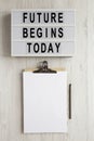 `Future begins today` words on a lightbox, clipboard with blank sheet of paper on a white wooden background, top view. Overhead,