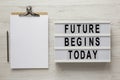 `Future begins today` words on a lightbox, clipboard with blank sheet of paper on a white wooden surface, top view. Overhead, fr