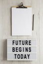 `Future begins today` words on a modern board, clipboard with blank sheet of paper on a white wooden surface, top view. Overhead